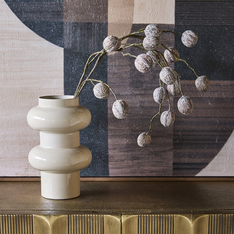 Libra Interiors Tiered Enamel Iron Vase in cream with stacked rounded tiers, holding a dried berry branch on a brass sideboard against abstract art.
