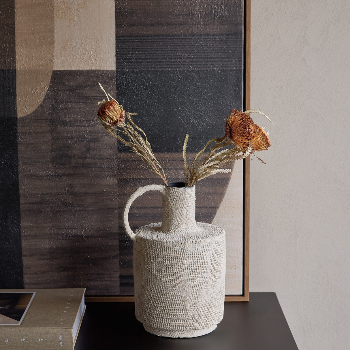 Libra Interiors Textured Cross-Hatch Handle Vase in cream holding dried banksia and grasses, styled on a dark console beside an abstract framed artwork.