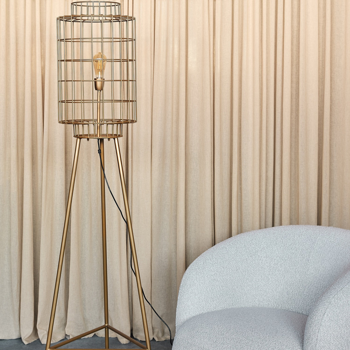 Libra Interiors Wire Cage Tripod Floor Lamp in brass with cylindrical iron cage shade and exposed bulb, beside a bouclé chair and beige curtains.