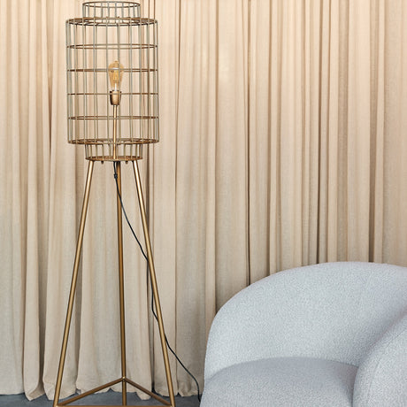 Libra Interiors Wire Cage Tripod Floor Lamp in brass with cylindrical iron cage shade and exposed bulb, beside a bouclé chair and beige curtains.