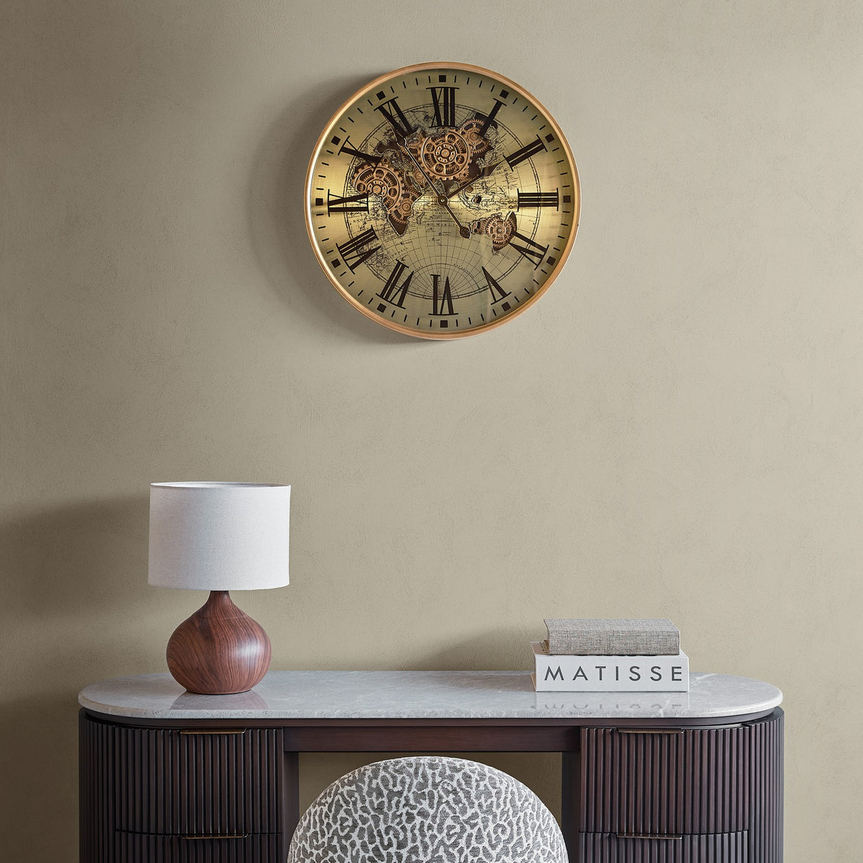 Libra Interiors Moving Cogs World Map wall clock in antique gold hung above a marble-topped console with a wooden lamp and books.