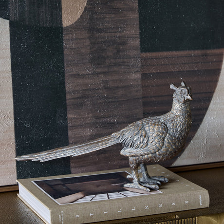 Libra Interiors Textured Antique Gold Pheasant Sculpture with long trailing tail and detailed plumage, displayed on a hardback book beside abstract artwork.