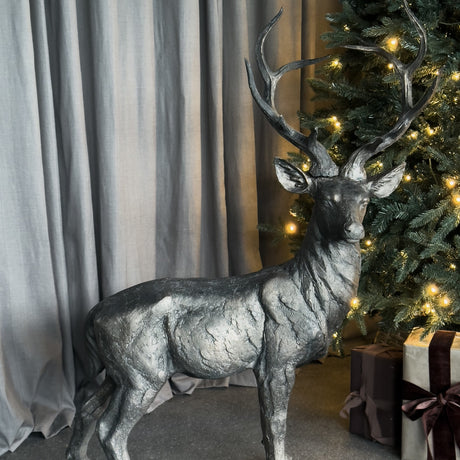 Libra Interiors Textured Bronze Standing Stag Sculpture with tall antlers, positioned beside a lit Christmas tree and wrapped gifts against grey curtains.