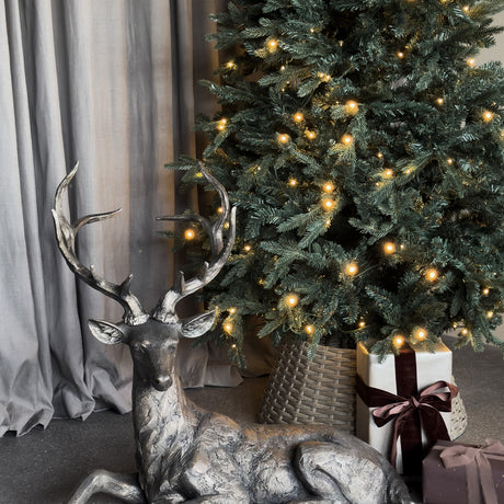 Libra Interiors Textured Resin Lying Stag Sculpture in bronze, resting with raised antlers beside a lit Christmas tree in a woven basket with wrapped gifts.