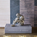 Libra Interiors Textured Resin Seated Male Sculpture in antique gold, head bowed over folded arms and knees, displayed on a grey Aesthetics of Design book.
