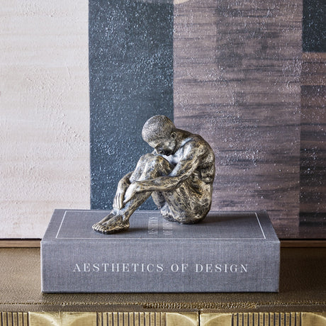 Libra Interiors Textured Resin Seated Male Sculpture in antique gold, head bowed over folded arms and knees, displayed on a grey Aesthetics of Design book.