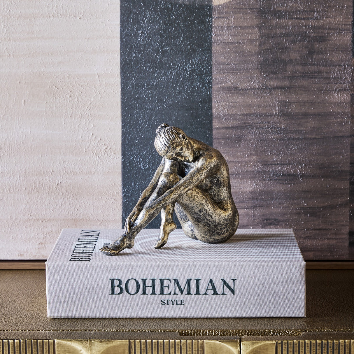 Libra Interiors textured resin seated female sculpture in antique gold, resting on a Bohemian Style book against an abstract painted backdrop.