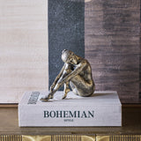 Libra Interiors textured resin seated female sculpture in antique gold, resting on a Bohemian Style book against an abstract painted backdrop.