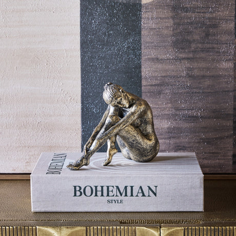 Libra Interiors textured resin seated female sculpture in antique gold, resting on a Bohemian Style book against an abstract painted backdrop.