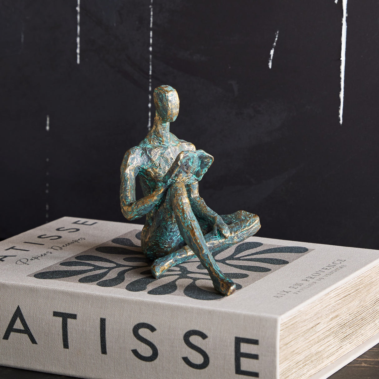 Libra Interiors Verdigris Seated Reading Man resin sculpture in antique gold and blue, displayed cross-legged atop a Matisse book against a dark backdrop.