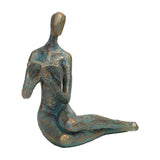 Libra Interiors Reading Woman verdigris resin sculpture in antique gold and blue, seated abstract figure leaning over an open book, on white.
