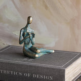 Libra Interiors Reading Woman resin sculpture in antique gold verdigris finish, seated with an open book, displayed on a grey design book.