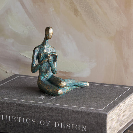 Libra Interiors Reading Woman resin sculpture in antique gold verdigris finish, seated with an open book, displayed on a grey design book.
