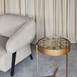 Libra Interiors Clock Top Round Antique Gold Side Table with glass clock face and cogs, placed beside a grey armchair against neutral curtains.