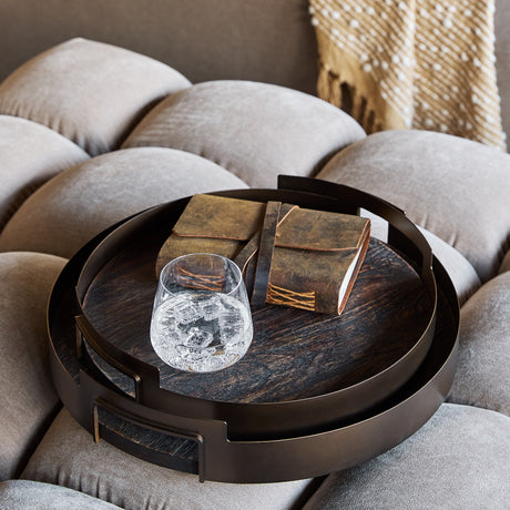 Libra Interiors Round Mango Wood and Smoked Gold nesting serving trays on a grey tufted sofa, holding a leather-bound journal and a glass of iced water.