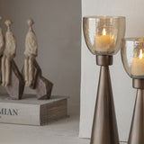 Libra Interiors Hammered Glass Smoke Gold Hurricane Lanterns with tapered iron bases and copper lustre glass cups holding lit candles, near a book.