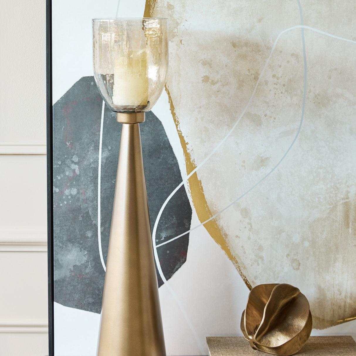 Libra Interiors Tapered Iron Hammered Glass Floor Lantern in gold with copper lustre glass vessel, styled beside abstract artwork in a contemporary living room.