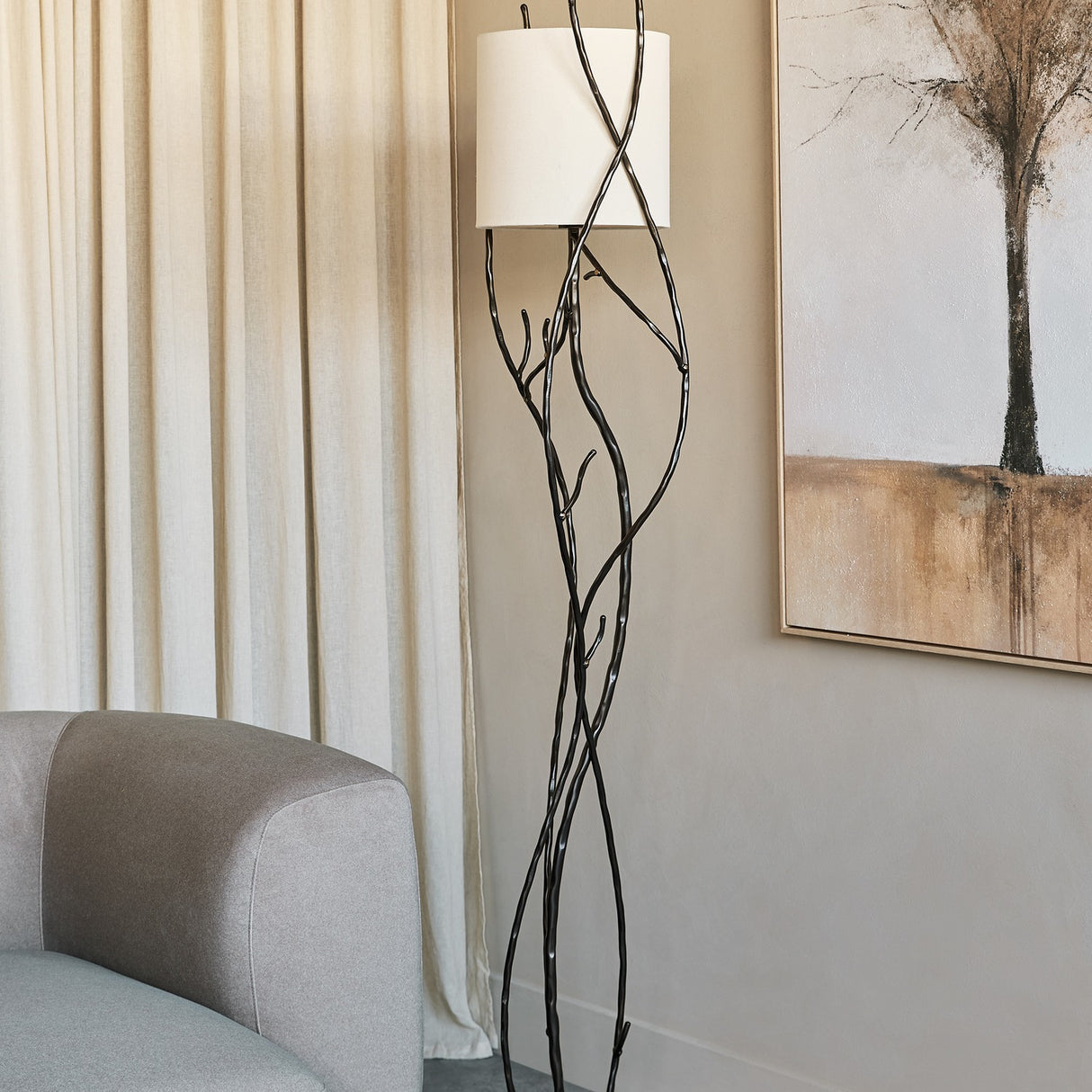 Libra Interiors Branch Iron Blackened Floor Lamp with twisted branch-like iron stem and round linen shade beside a grey sofa and cream curtains.