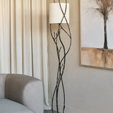 Libra Interiors Branch Iron Blackened Floor Lamp with twisted branch-like iron stem and round linen shade beside a grey sofa and cream curtains.