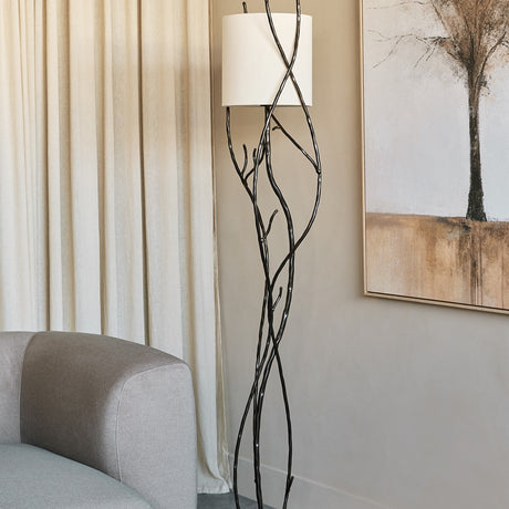 Libra Interiors Branch Iron Blackened Floor Lamp with twisted branch-like iron stem and round linen shade beside a grey sofa and cream curtains.
