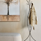 Libra Interiors branch-style iron tripod coat stand in black with bronze highlights, holding a beige jacket in a hallway beside a framed tree artwork.