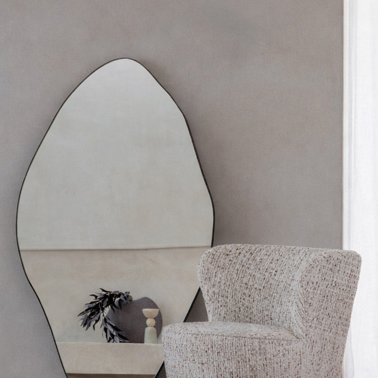 Libra Interiors Corrugated Iron Organic Wall Mirror with pebble-like silhouette and gilded oak frame, hung above a boucle armchair in a neutral living room.