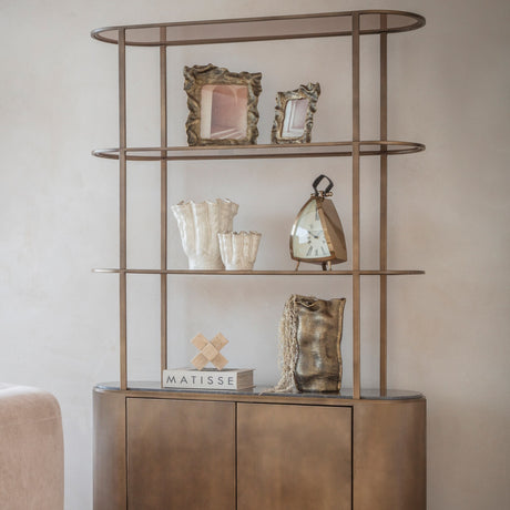 Libra Interiors Marble Top Corrugated Metal Wall Shelf in gold with three tiers displaying frames, vases, a clock and decor above a ribbed cabinet.