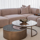 Libra Interiors Marble & Glass Round Nesting Coffee Table in gold with tinted glass top and grey marble drum base, styled beside a taupe sofa.