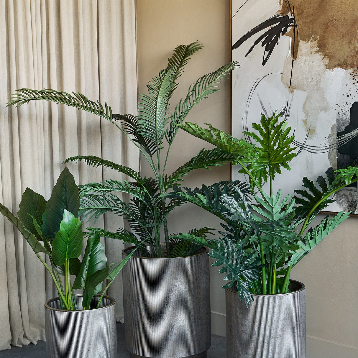 Libra Interiors Corrugated Iron Ridged Floor Planters in champagne gold, three ridged cylindrical sizes holding tall palms and leafy plants in a neutral room.