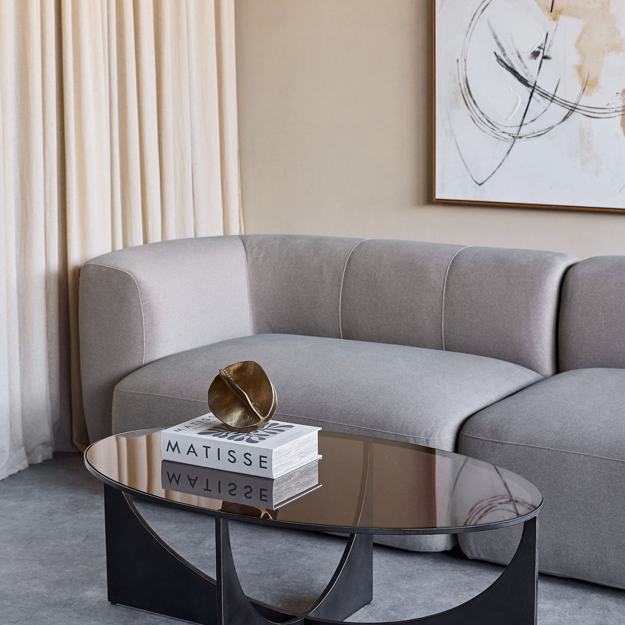 Libra Interiors Bronze Glass Sculptural Coffee Table in black with oval tinted top and curved iron base, styled with Matisse books beside a grey sofa.