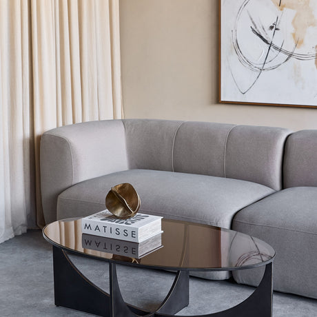 Libra Interiors Bronze Glass Sculptural Coffee Table in black with oval tinted top and curved iron base, styled with Matisse books beside a grey sofa.