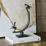 Libra Interiors Rough Cast Two Figure Balance Sculpture in bronze, with stylised figures on a curved arc, displayed on a book beside abstract art.