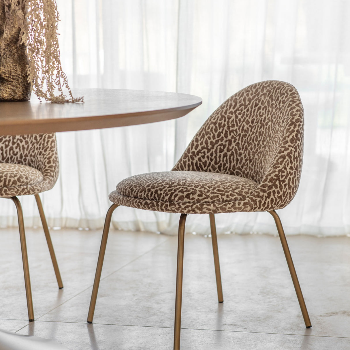 Libra Interiors Woven Animal Print dining chair in beige and taupe jacquard fabric on slim antique gold brass legs, beside a round wooden table.