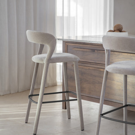 Libra Interiors Velvet Wrapped Curved Back Bar Stools in taupe with fabric-covered tapered legs and black footrests, set at a wooden kitchen island.