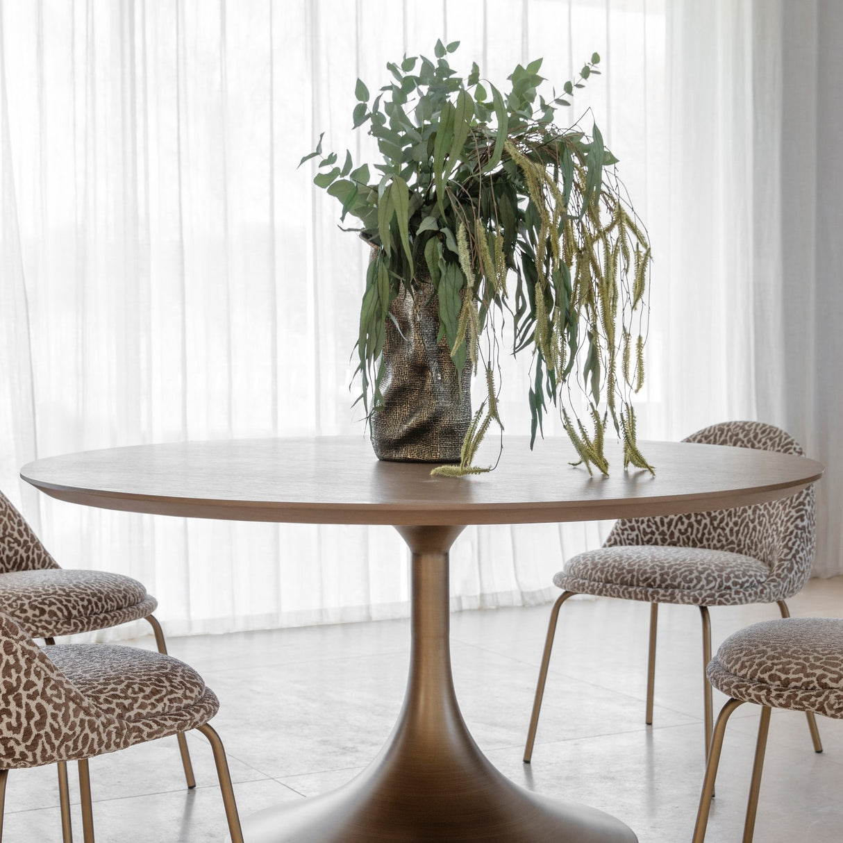 Libra Interiors Brushed Brass Pedestal Round Dining Table with light wood top and flared antique brass base, styled with a leafy plant and patterned chairs.