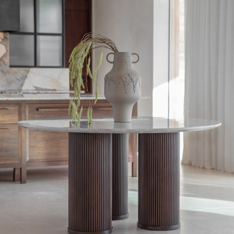 Libra Interiors Fluted Marble Top Round Dining Table in grey with three walnut fluted legs, styled with a stone vase and trailing foliage in a dining room.