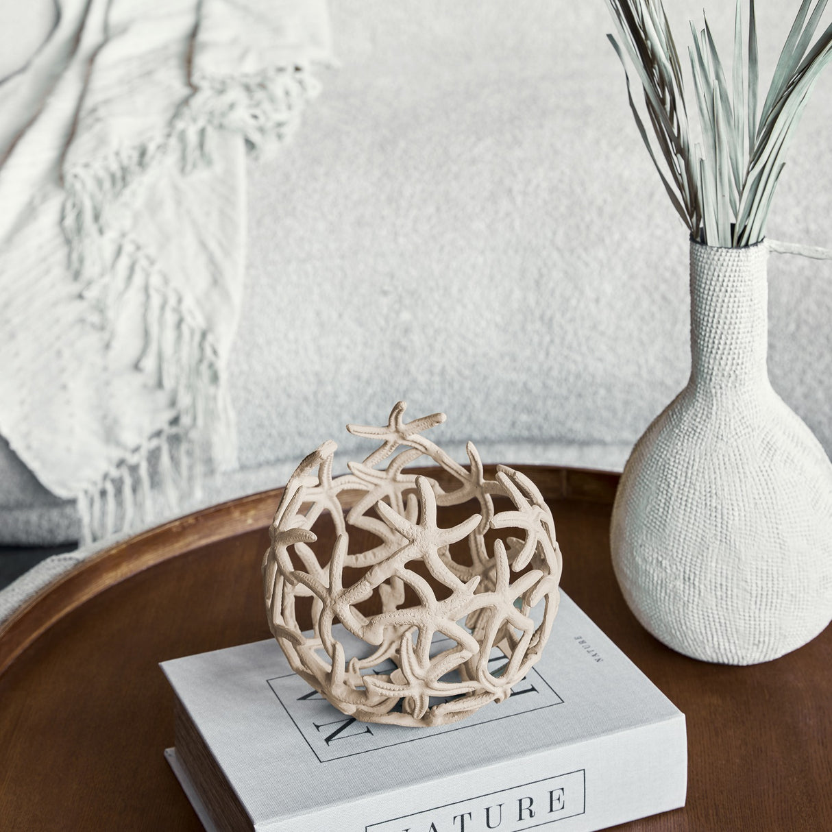 Libra Interiors Openwork Starfish Aluminium Decorative Bowl in beige, styled on a book atop a wooden tray beside a textured white vase with foliage.