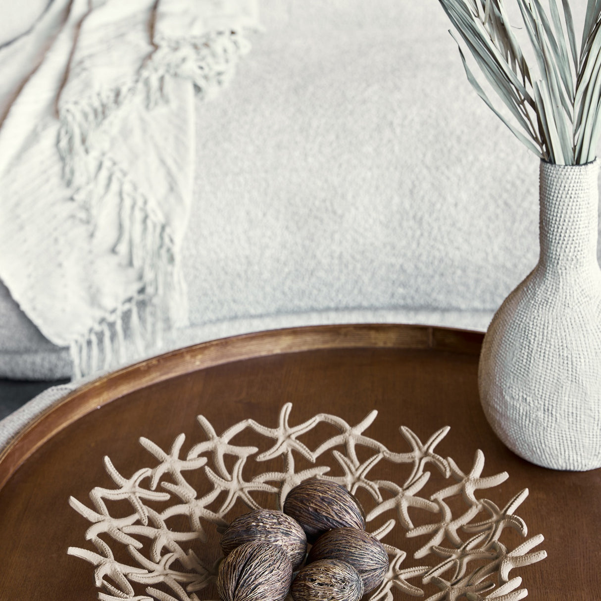 Libra Interiors Starfish Openwork Shallow Bowl in beige aluminium on a wooden tray, holding dried pods beside a textured vase and throw in a coastal setting.