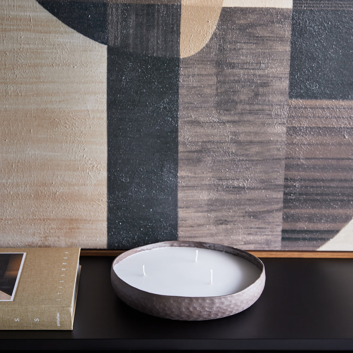Libra Interiors hammered aluminium three-wick bowl candle in brown with white wax, on a dark console beside a book and abstract artwork.
