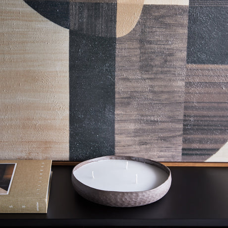 Libra Interiors hammered aluminium three-wick bowl candle in brown with white wax, on a dark console beside a book and abstract artwork.