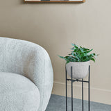 Libra Interiors hammered white aluminium planter with leafy green foliage on a slim black four-leg stand, beside a bouclé armchair in a neutral living room.