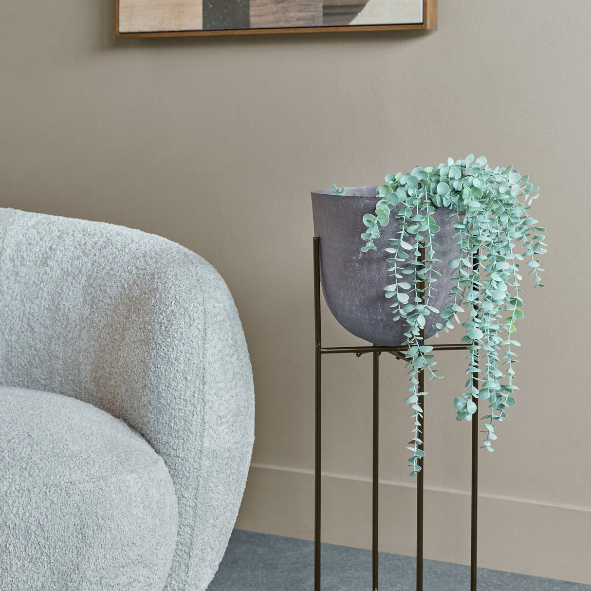 Libra Interiors hammered aluminium raised planter in brown with trailing eucalyptus, on a black iron stand beside a grey bouclé chair in a living room.