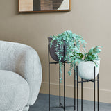 Libra Interiors hammered aluminium raised planter in brown with trailing eucalyptus, beside a white planter on stand and boucle armchair in a living room.
