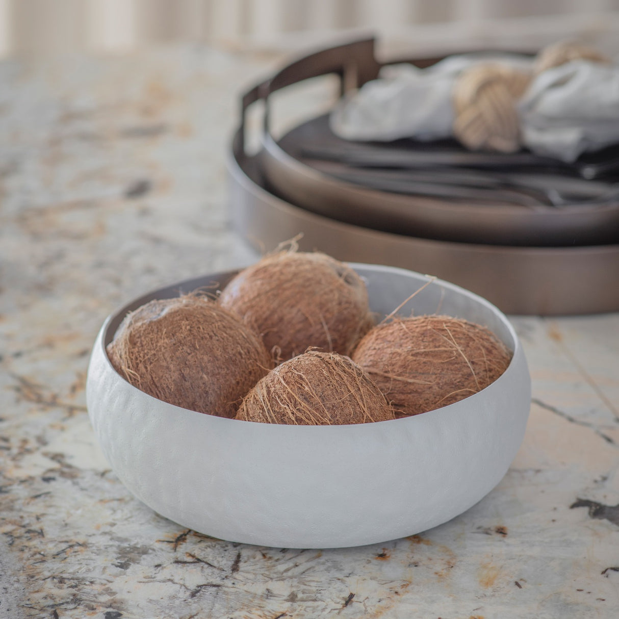 Libra Interiors Hammered Stonewash Aluminium Bowl in white holding whole coconuts on a marble surface, with a dark round tray behind.