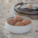 Libra Interiors Hammered Stonewash Aluminium Bowl in white holding whole coconuts on a marble surface, with a dark round tray behind.