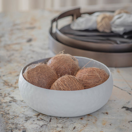 Libra Interiors Hammered Stonewash Aluminium Bowl in white holding whole coconuts on a marble surface, with a dark round tray behind.