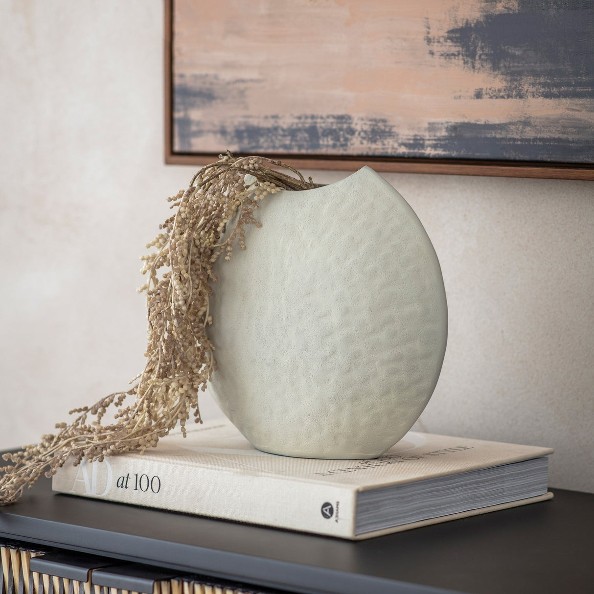 Libra Interiors Hammered White Stonewash Moon Vase holding dried stems, displayed on an AD at 100 book atop a dark sideboard beneath abstract art.