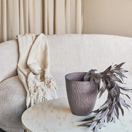 Libra Interiors hammered stonewash aluminium tapered planter in brown on a light mosaic coffee table, holding dark foliage beside a bouclé sofa and throw.