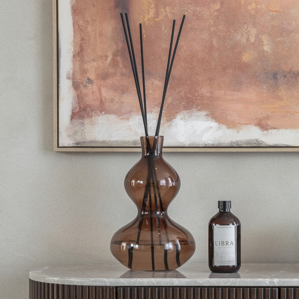 Libra Interiors Sculptural Glass Reed Diffuser in brown, curved double-gourd vessel with black reeds, on a marble console beneath an abstract artwork.