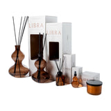 Libra Interiors Sculptural Glass Reed Diffuser in brown, shown in two sizes with black reeds beside white Palo Santo packaging, spray and candle on white.
